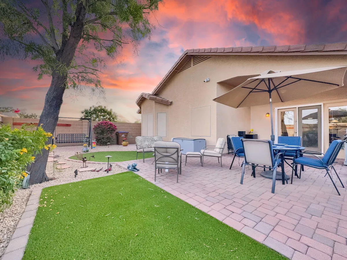 An inviting patio space with comfortable furniture under a large umbrella, set against a backdrop of a landscaped garden and open sky. The mood is serene and perfect for morning coffee or sunset relaxation.