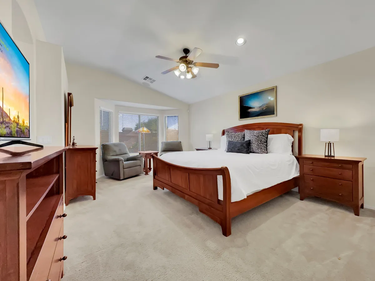 A spacious master suite featuring a king-sized bed with plush linens, a mounted television, and a cozy sitting area with upholstered chairs. Ample natural light streams in through large windows, creating a calming ambiance ideal for relaxation and restful nights.