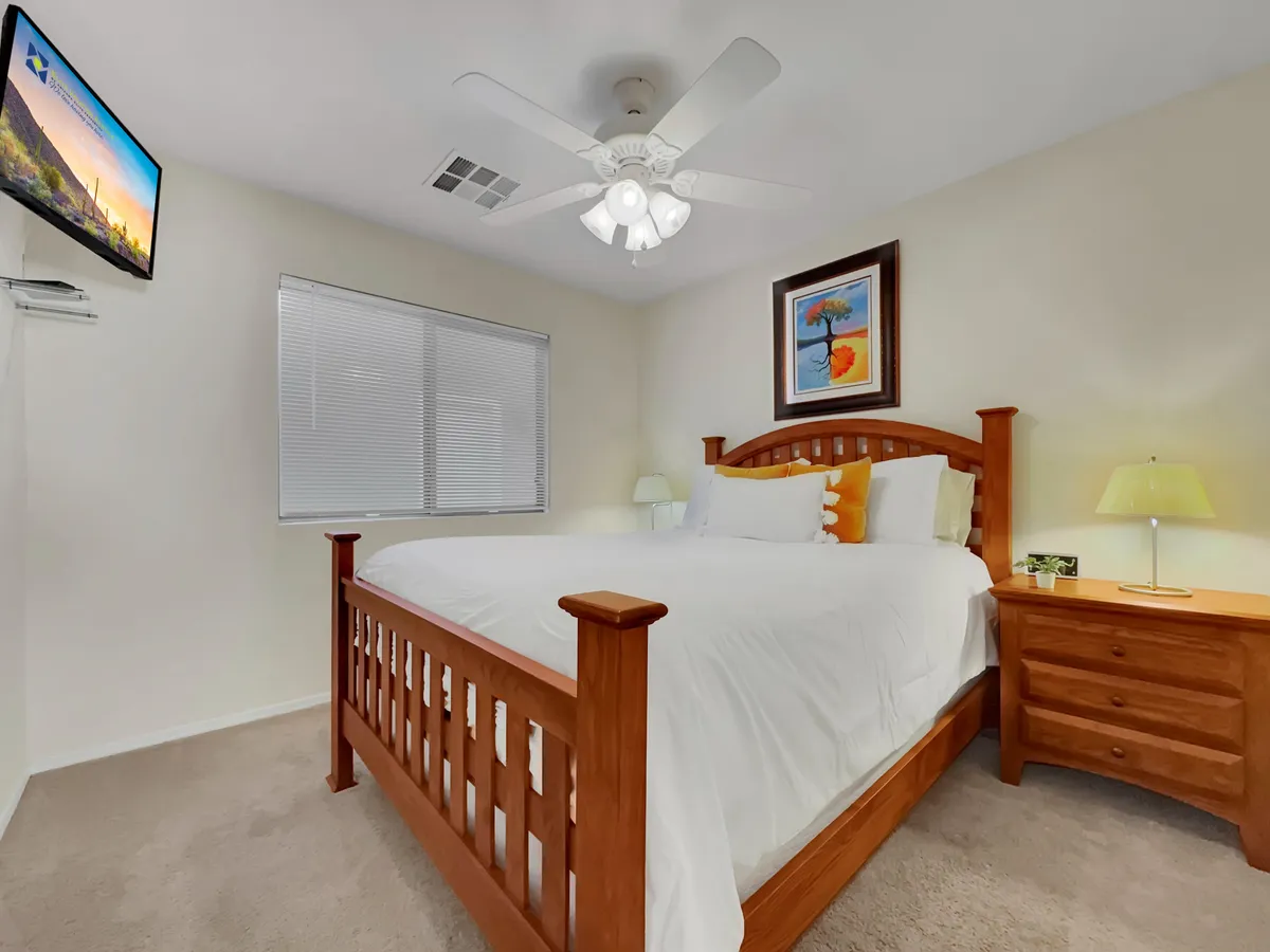 A welcoming guest bedroom with a queen-sized bed, tasteful decorative pillows, framed artwork, a wooden nightstand, and a wall-mounted television. Sunlight illuminates the soft textures, making the room feel both inviting and restful.