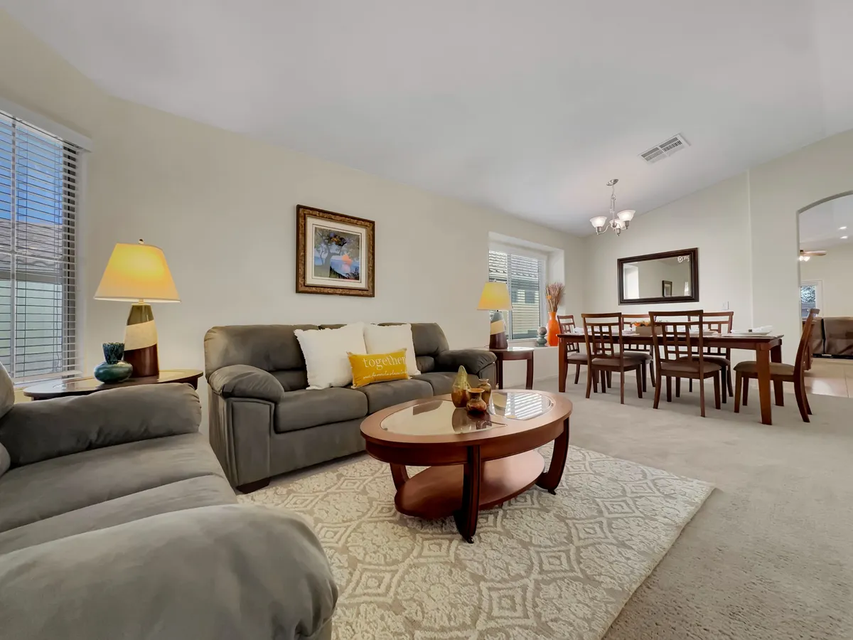 A bright, open-concept area combining a sofa, coffee table, and adjacent dining space, punctuated by decorative artwork and a lamp. The setting feels homey and versatile, suitable for both group gatherings and quiet moments, while large windows ensure plenty of daylight.
