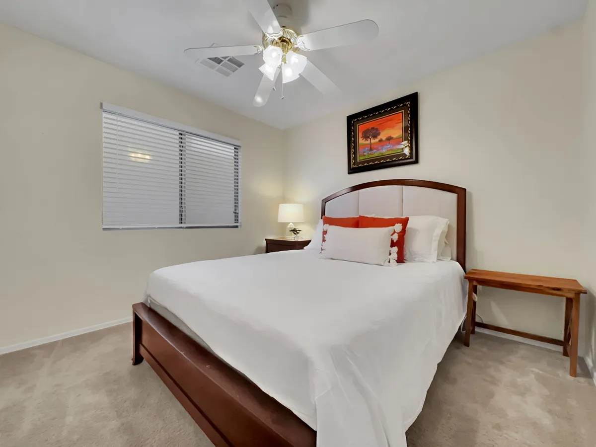 A serene bedroom with white linens, decorative pillows, a bedside lamp, framed artwork above the bed, and a wooden side table. The room is bathed in natural light, offering a peaceful place for relaxation or a good night's sleep.