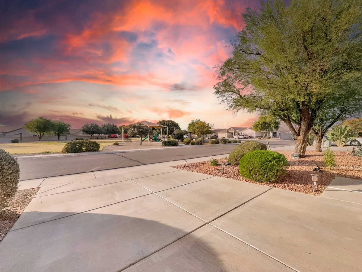 This image captures the neatly landscaped front yard of the home with lush green trees, a smooth concrete driveway, and a vibrant sunset sky overhead. The setting is serene and inviting, suggesting a comfortable space for evening activities and outdoor enjoyment.