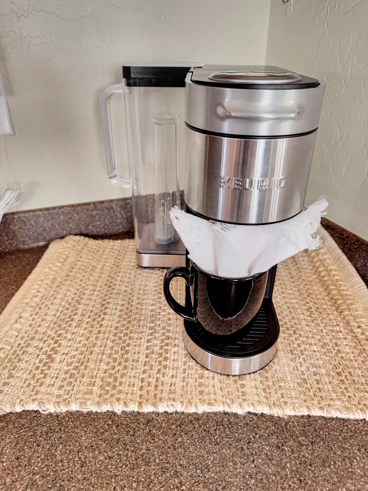 Close-up of a Keurig coffee maker with a black mug on a woven placemat, evoking a sense of daily rituals and relaxation. The cozy arrangement highlights the comfort and convenience provided in the kitchen.