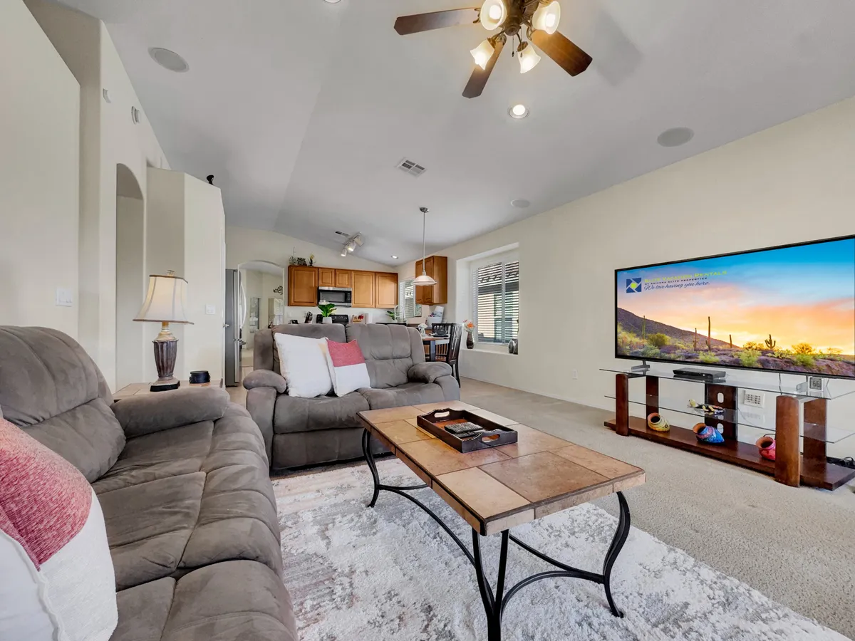 A spacious living area featuring comfortable sofas, a central coffee table, a large television, and a view toward an open kitchen. The room is softly lit and feels welcoming, ideal for family movie nights or group relaxation. The setup encourages both entertainment and conversation in a relaxed setting.