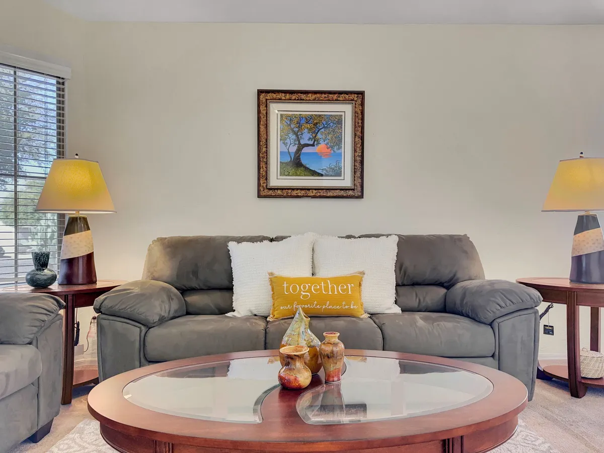 A cozy living space with a plush sofa adorned with decorative pillows, a coffee table, and vibrant wall art. The room is bathed in natural light and offers a glimpse of greenery outside, suggesting garden views. The inviting arrangement is ideal for socializing, reading, or relaxing with loved ones—including furry friends.