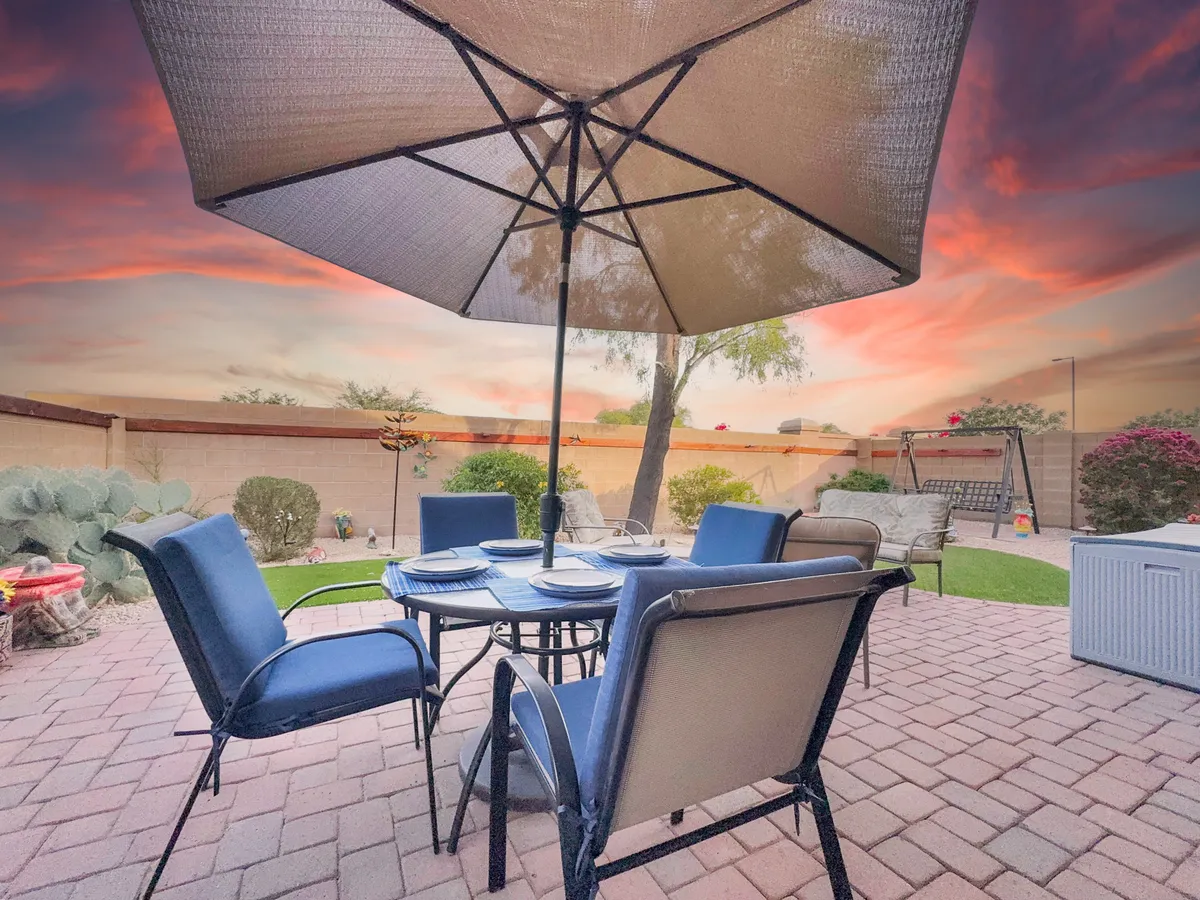 A beautifully landscaped backyard featuring a table with chairs beneath a large umbrella, surrounded by lush greenery and outdoor furniture. The space feels inviting for both social gatherings and peaceful relaxation, with ample room for children and pets to play.